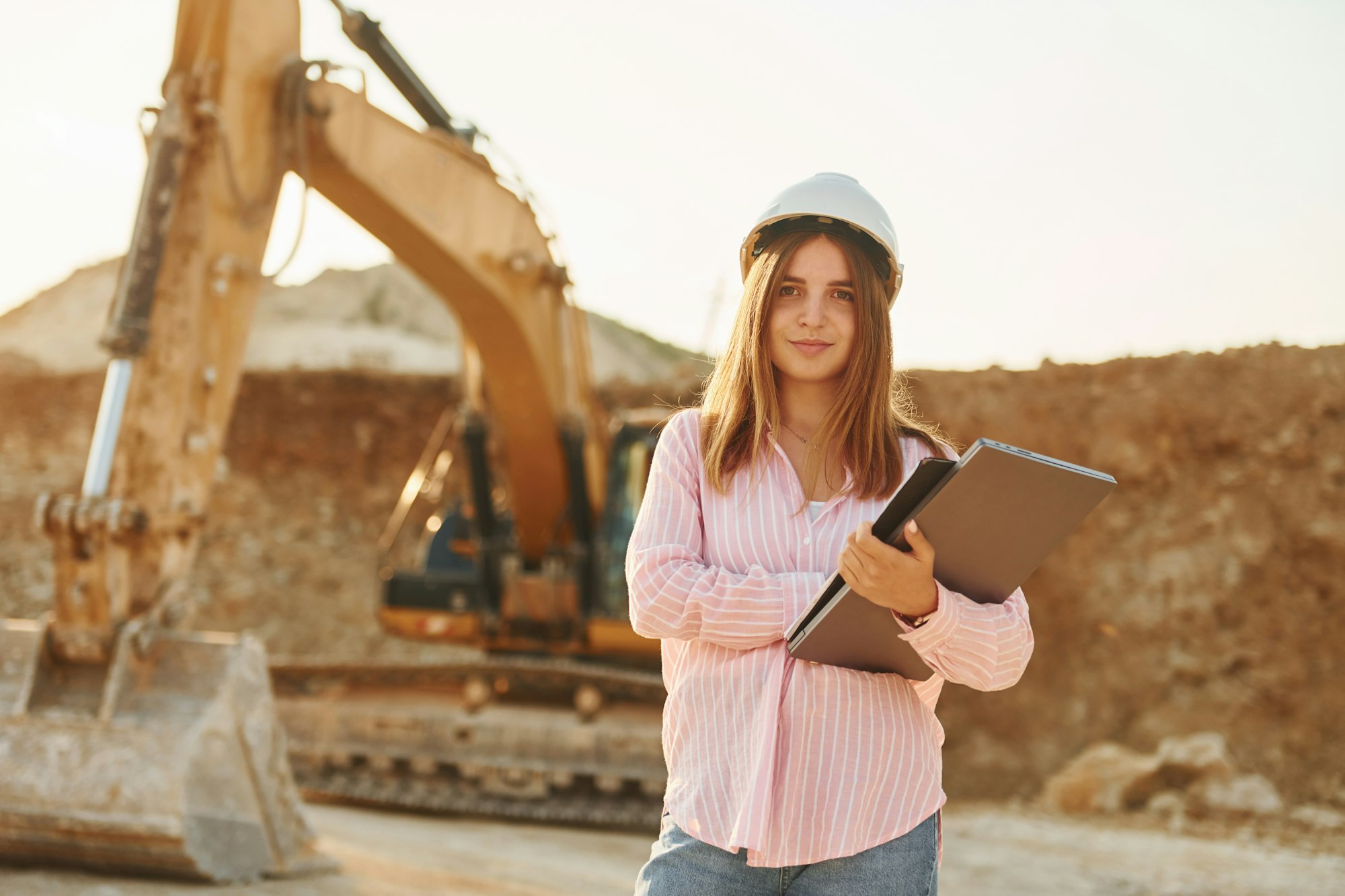 With documents. Young woman with documents in hard hat is standing on the borrow pit